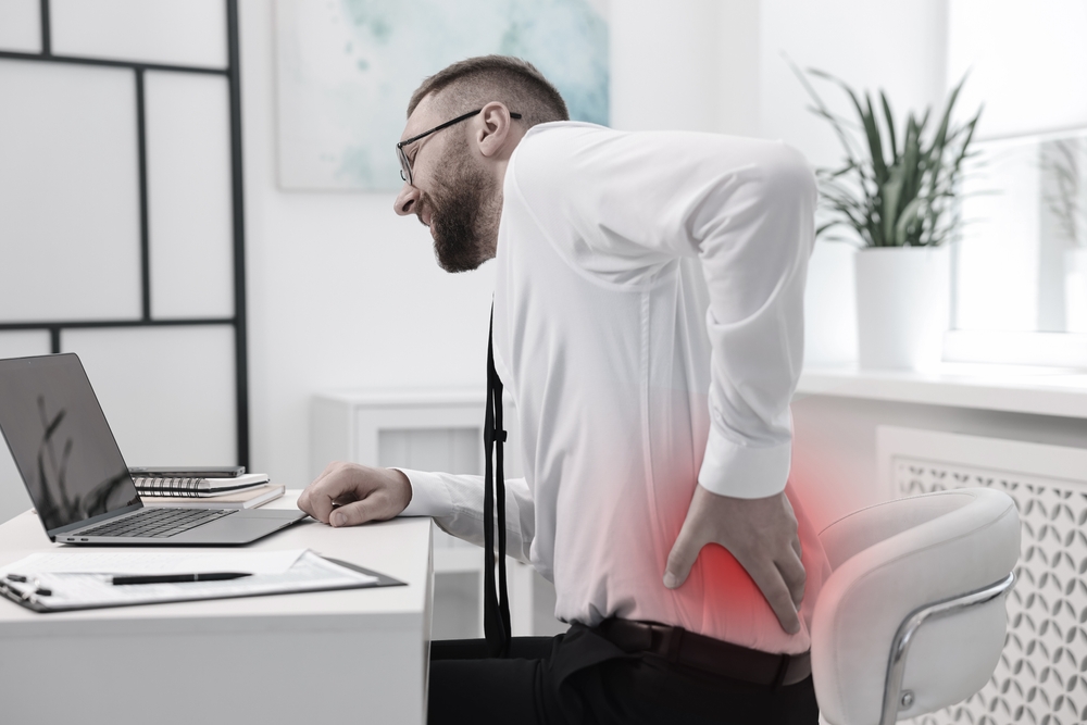 Bearded man in white shirt standing at desk, bending backward while holding his lower back with red highlighted pain area, suffering from acute back strain during work on laptop
